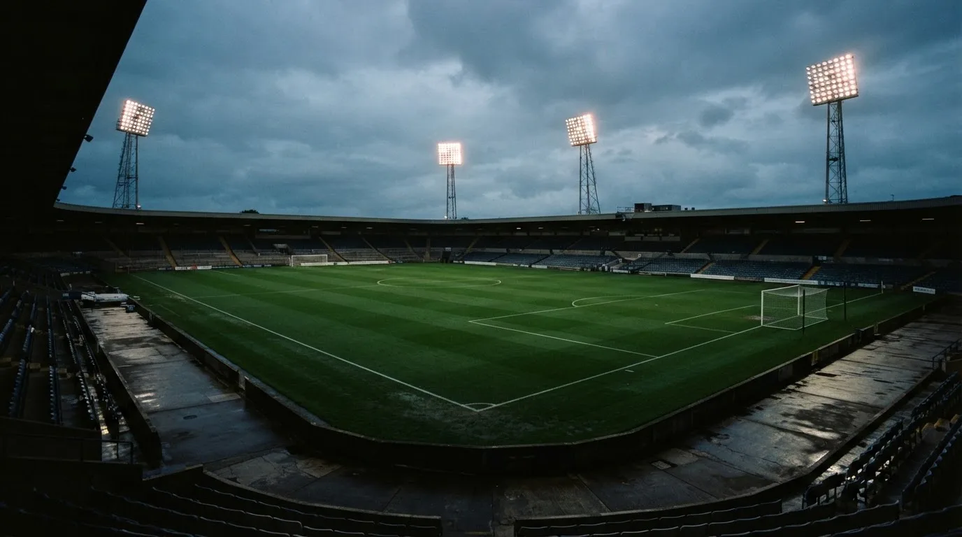 Ein Fußballplatz mit Flutlichtmasten in der Abenddämmerung, aus der Ferne gesehen, leere Tribünen und nasser Rasen unter bewölktem Himmel.