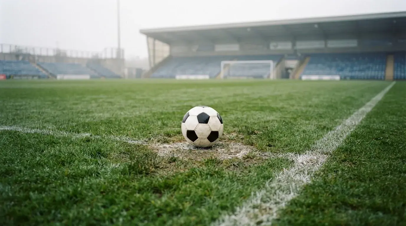 Ein Fußball liegt auf dem Elfmeterpunkt eines leeren Rasenplatzes, im Hintergrund eine Tribüne im Dunst.