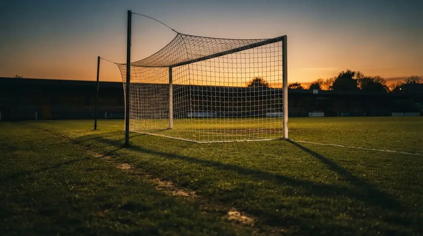 Ein Fußballtor mit Netz bei Sonnenuntergang auf einem leeren Platz, die Schatten der Torpfosten liegen lang auf dem Gras.