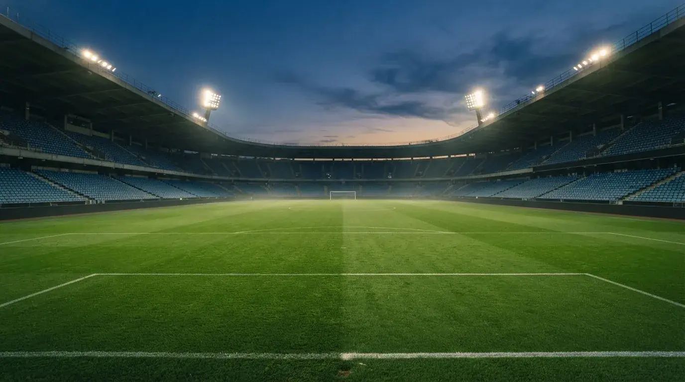 Eine leere Stadiontribüne unter Flutlicht aus der Ferne betrachtet, die Rasenfläche leuchtet grün in der Dämmerung.