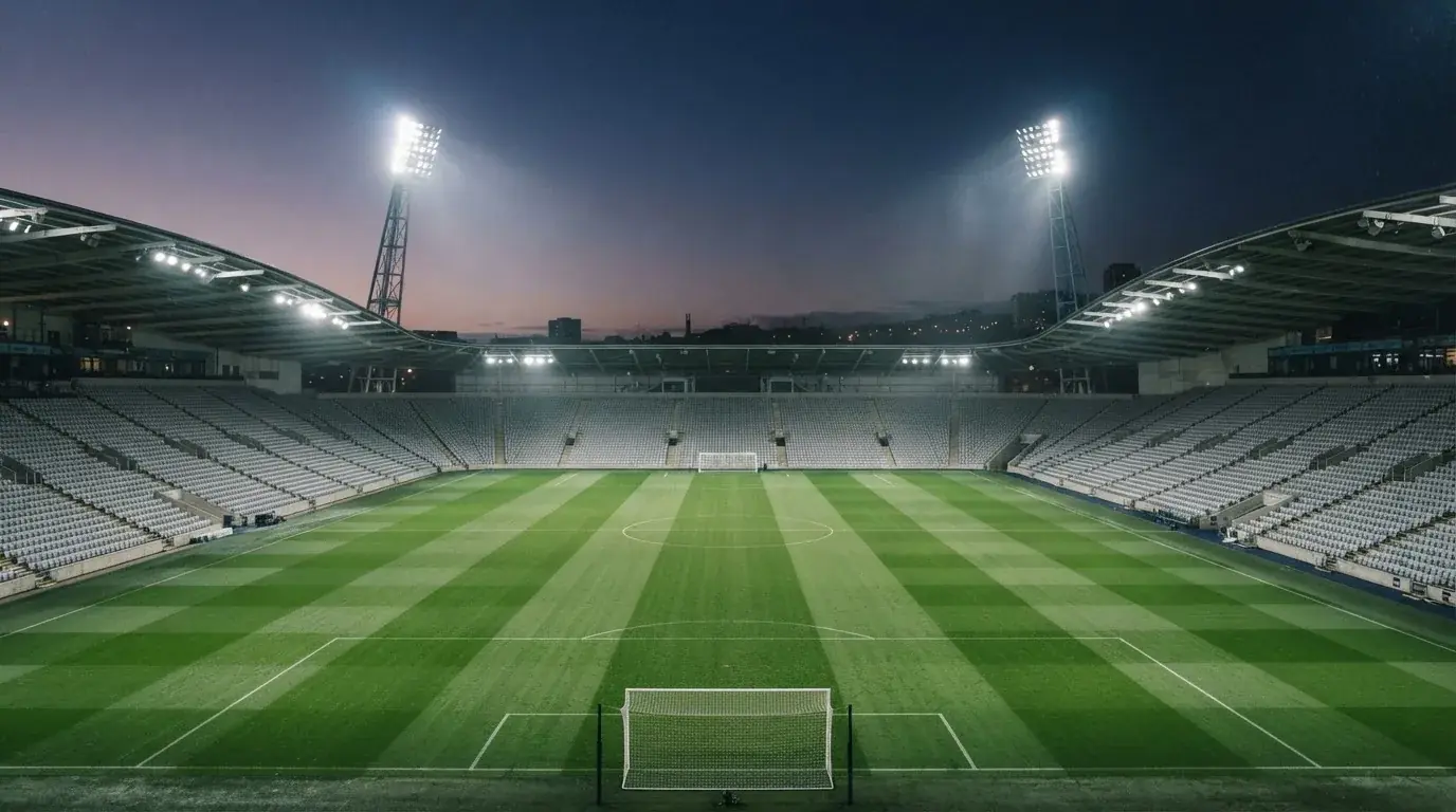 Eine leere Stadiontribüne unter Flutlicht aus der Ferne betrachtet, die Rasenfläche leuchtet grün in der Dämmerung.