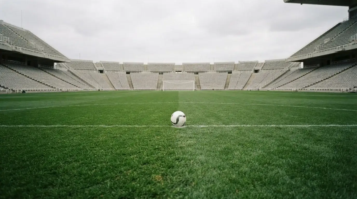 Ein Fußball liegt auf dem Anstoßpunkt eines leeren Rasenplatzes, im Hintergrund eine ruhige Tribüne unter bedecktem Himmel.