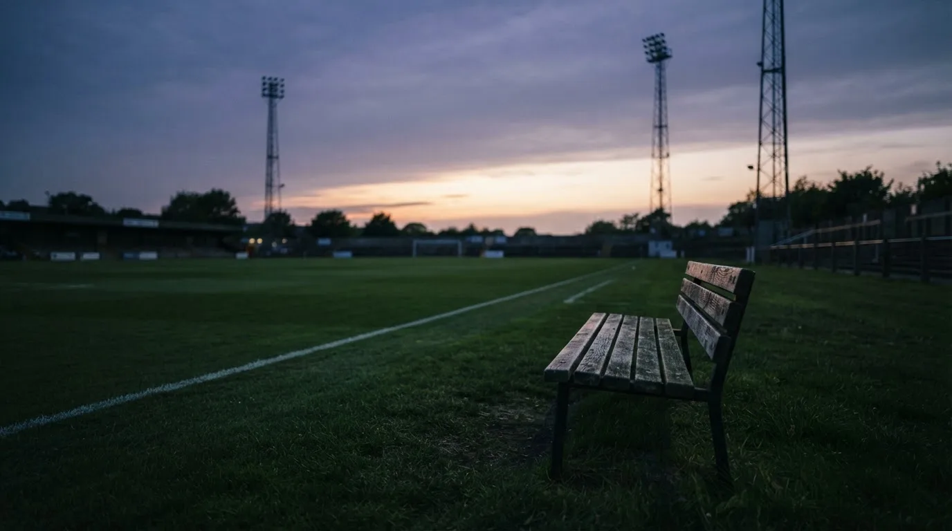 Eine leere Parkbank am Rand eines Fußballfelds in der Abenddämmerung, im Hintergrund ragen dunkle Flutlichtmasten in den Himmel.